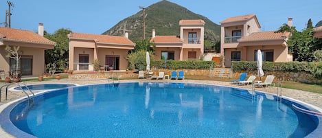 A swimming pool surrounded by a building with a tiled roof, a mountain in the background, and a clear sky.