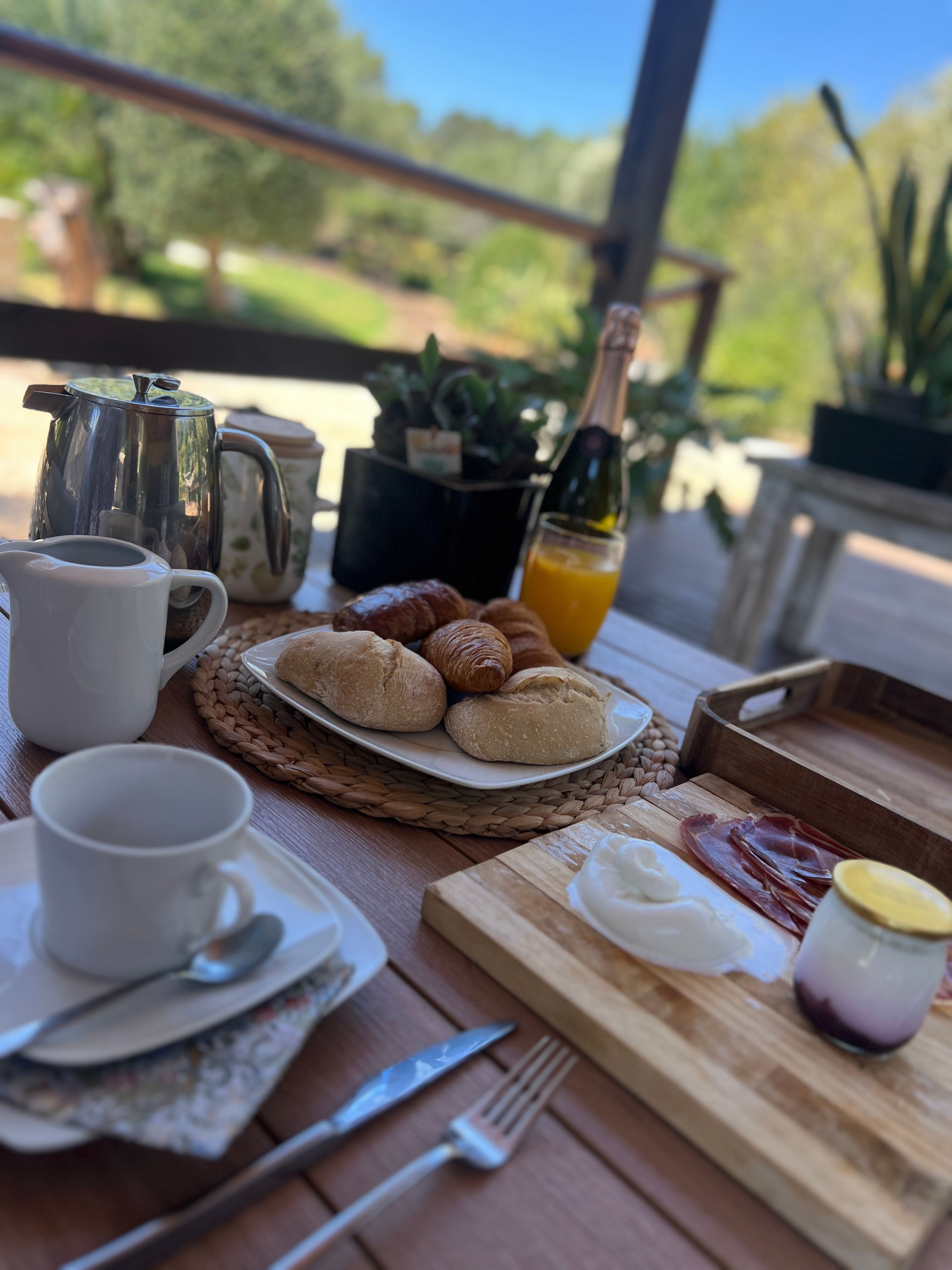 Une table en bois dressée pour le petit-déjeuner, avec une cafetière, des croissants, une bouteille de jus de fruits et une assiette de nourriture.