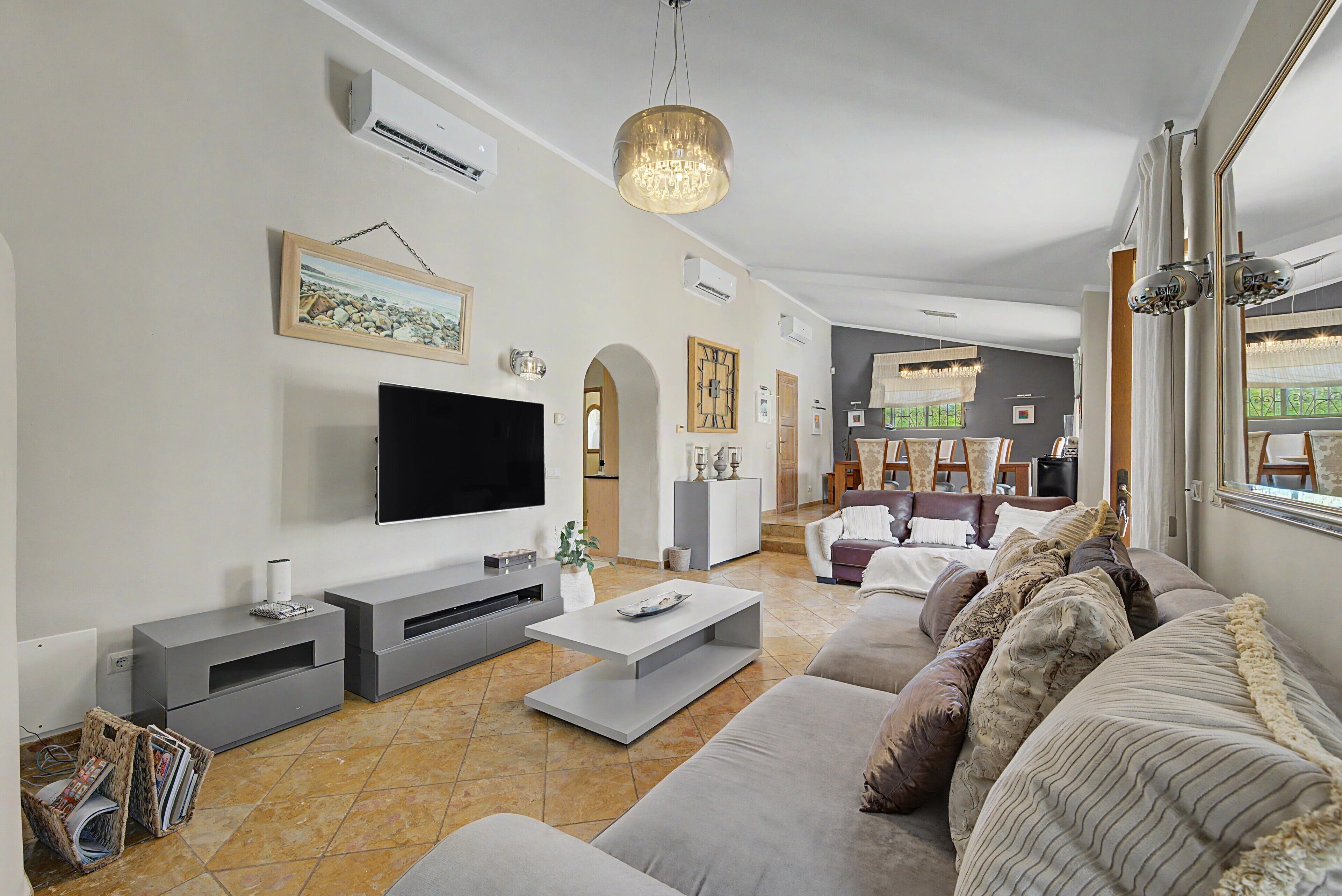 A modern living room with a flat-screen TV, grey sofas, a coffee table, and a chandelier.