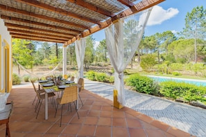 Ein Essbereich im Freien mit einem Tisch für vier Personen, umgeben von einer Pergola mit einem Reetdach, einem Pool und einem Garten mit verschiedenen Pflanzen und Bäumen.