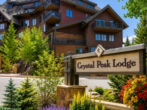 A sign for Crystal Peak Lodge with a stone base, surrounded by greenery and a multi-story building in the background. - Crystal Peak Lodge 7403 ~ Ski-in/Ski-out ~ NEW LISTING ~ By CoralTree Residences (Breckenridge)