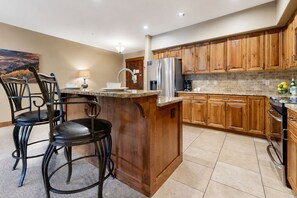 A kitchen with wooden cabinets, a stainless steel refrigerator, a marble countertop, and a dining area with two chairs. - Crystal Peak Lodge 7403 ~ Ski-in/Ski-out ~ NEW LISTING ~ By CoralTree Residences (Breckenridge)