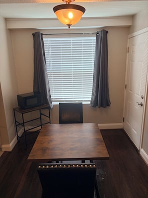 Un comedor con una mesa de madera, sillas, un estante pequeño con un microondas y una ventana con persianas y cortinas.