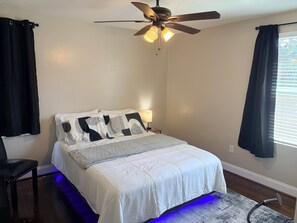 Un dormitorio con cama, ventilador de techo, mesita de noche y ventana con cortinas.