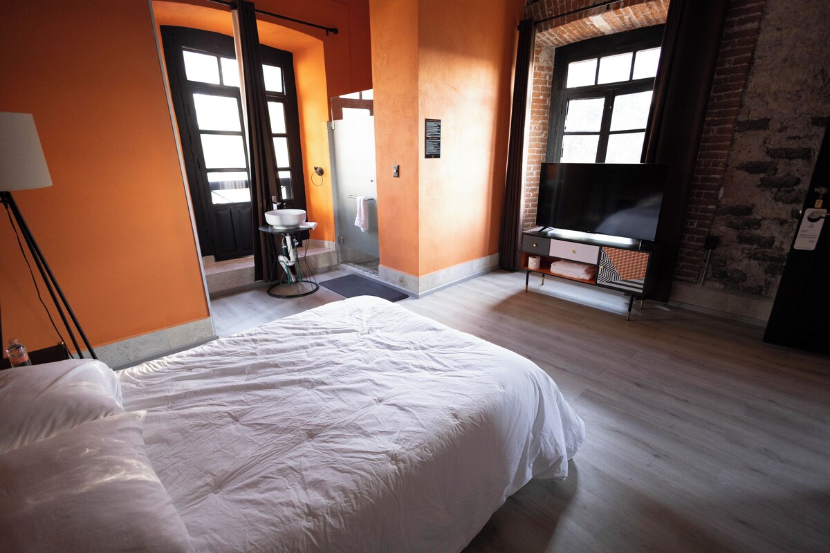 A hotel room with a large bed, a television, a bathroom with a sink and mirror, and a brick wall.