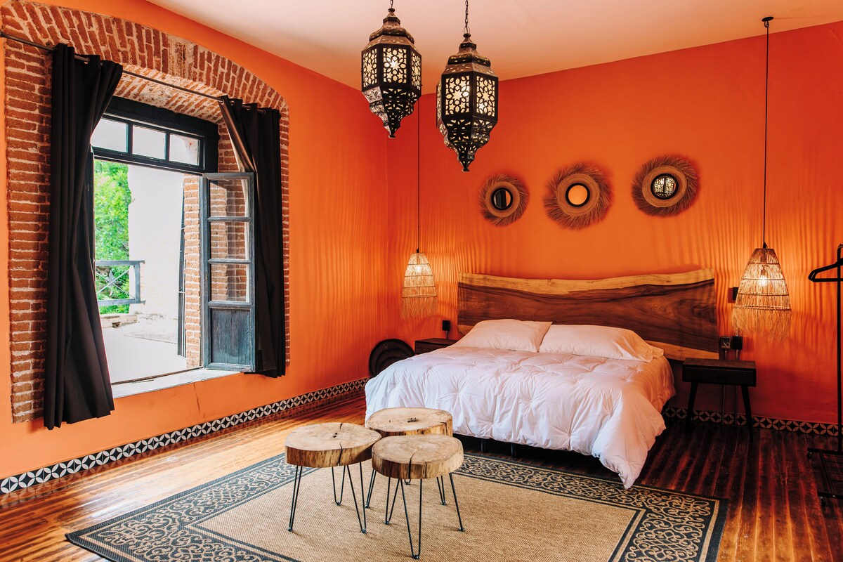 A room with a large bed, two hanging lanterns, a window with curtains, and a patterned rug.