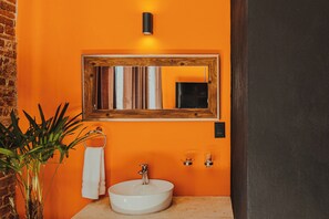 A bathroom with an orange wall, a wooden-framed mirror, a white sink, and a potted plant. - Casa Grande Rio (Rioverde)