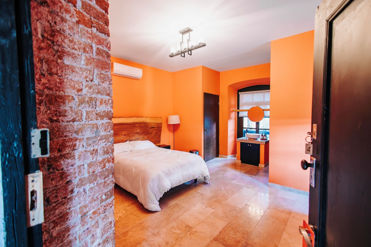A hotel room with a brick wall, a bed, a nightstand, a lamp, a window, and a bathroom visible through an archway.