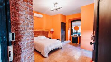 A hotel room with a brick wall, a bed, a nightstand, a lamp, a bathroom with a sink, and a door.