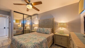 A bedroom with a bed, bedside tables, lamps, a ceiling fan, and a mirror. - Modern and Bright Kahana Retreat, a Cozy Coastal 1BR with Pool & AC, Ocean-View (Lahaina)