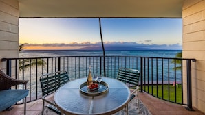 A balcony with a table set for two, overlooking a body of water with a distant mountain range. - Modern and Bright Kahana Retreat, a Cozy Coastal 1BR with Pool & AC, Ocean-View (Lahaina)