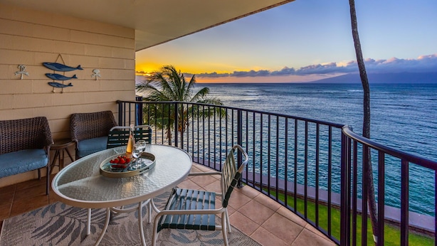 A balcony with a table, chairs, and a view of the ocean at sunset. - Modern and Bright Kahana Retreat, a Cozy Coastal 1BR with Pool & AC, Ocean-View (Lahaina)