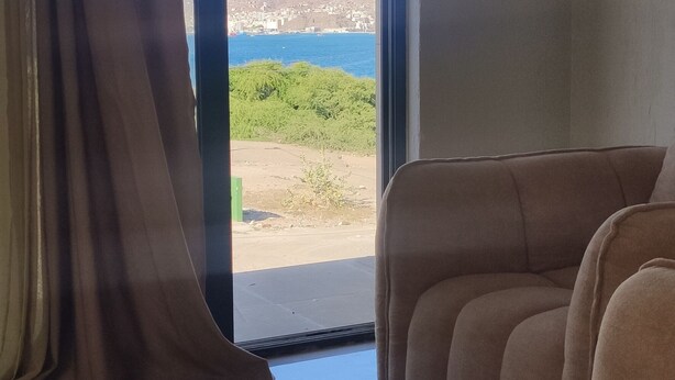 Habitació amb finestra gran, sofà i vista a un paisatge costaner.