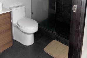 Een badkamer met een wit toilet, een houten kastje, een douchecabine met glazen wanden en een bruin badmat.