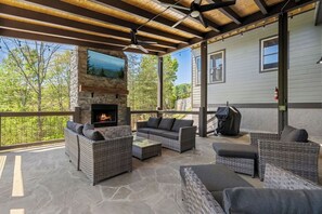 An outdoor patio with stone flooring, wicker furniture, a stone fireplace, and a TV mounted on the wall. - New | Mtn Views & Modern Luxury | Sleeps 12 | 5 Fireplaces | Pool & Hot Tub (Sevierville)