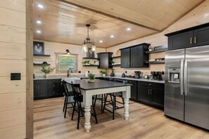 A modern kitchen with a central island, black cabinetry, stainless steel appliances, and wooden flooring. - New | Mtn Views & Modern Luxury | Sleeps 12 | 5 Fireplaces | Pool & Hot Tub (Sevierville)