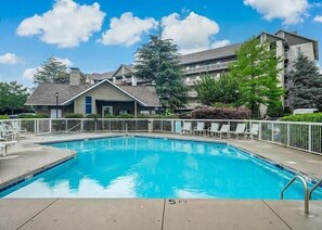 A swimming pool with a white fence, a small building with a blue door, and a large multi-story building in the background. - Whispering Pines 634 | 4BR Luxury Condo in the Heart of Pigeon Forge Sleeps 10 (Pigeon Forge)