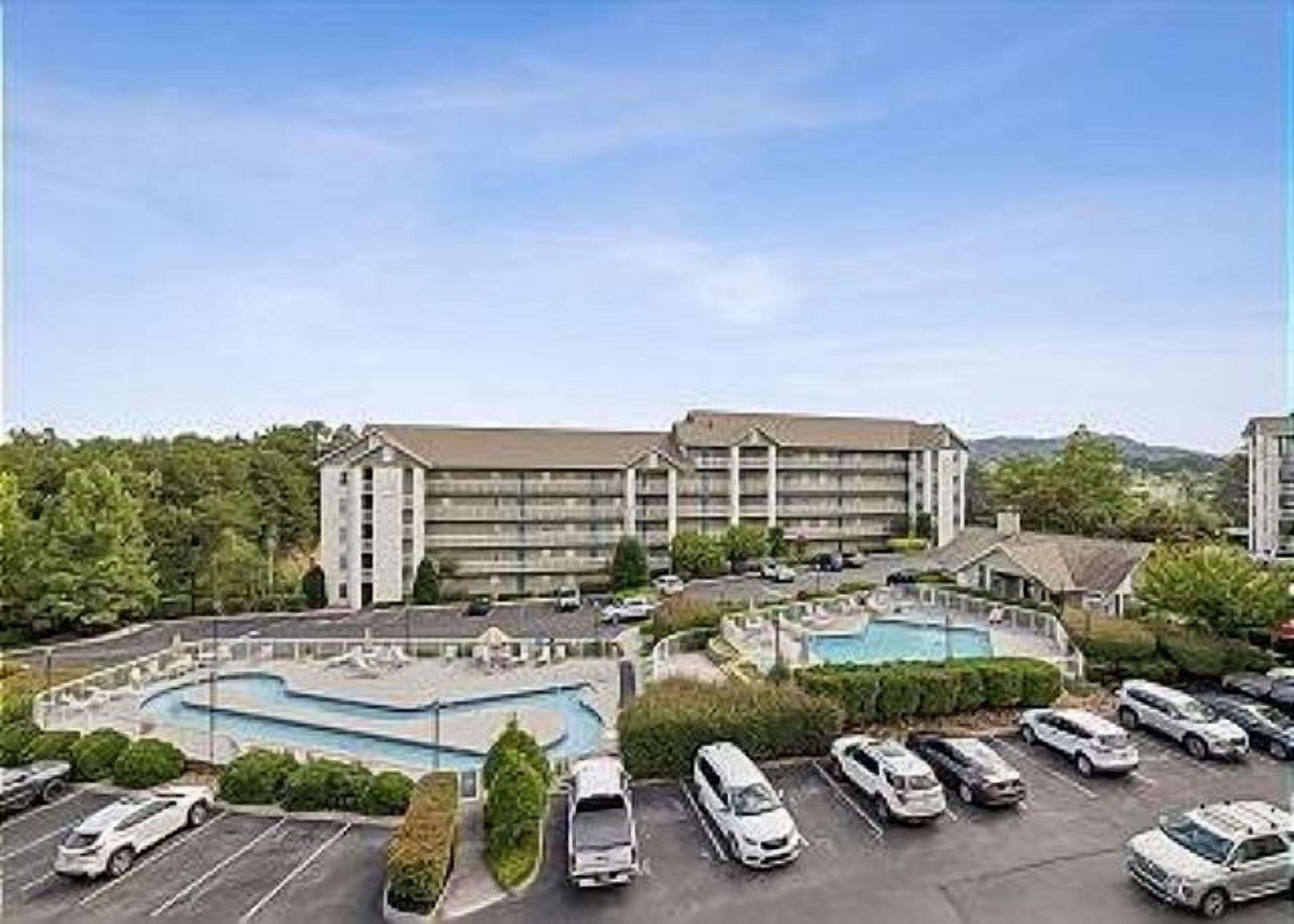 Un hotel con piscina, estacionamiento y montañas al fondo.