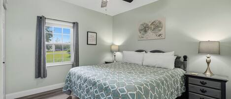 A bedroom with a bed, a nightstand, a lamp, a ceiling fan, a window with curtains, and a wall with a framed picture.