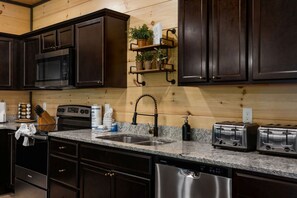 A kitchen with dark wood cabinets, stainless steel appliances, granite countertops, a built-in oven, a dishwasher, and a wooden wall with a shelf holding plants and a sign. - 12 Bedroom|Brand New|Pool|Games|Movie Room|Free Tkts (Sevierville)