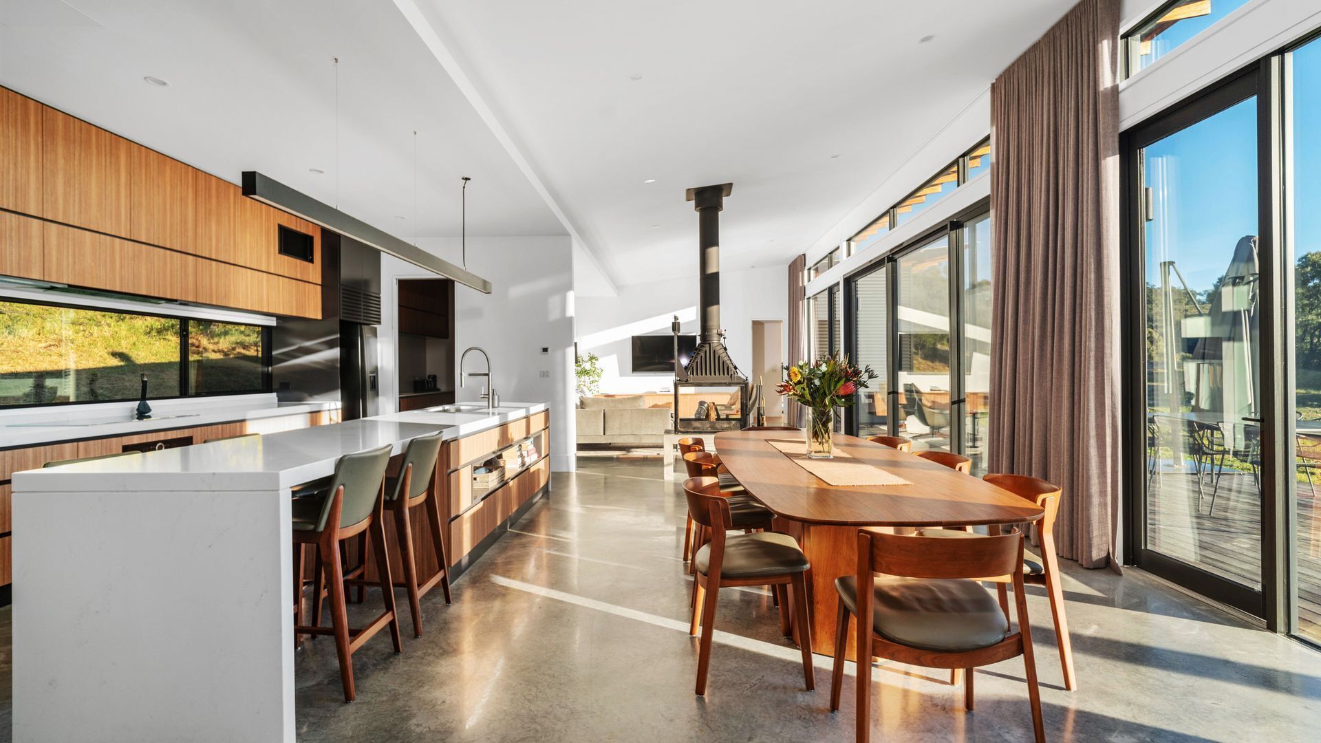 A modern kitchen with a central island, a dining area with a large table, and a view of the outdoors through large windows.