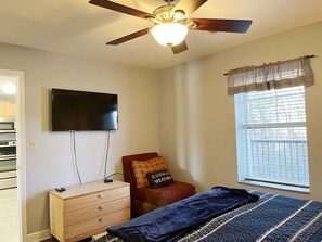 Ein Schlafzimmer mit einem Bett, einem Schminktisch, einem an der Wand befestigten Fernseher, einem Deckenventilator und einem Fenster mit Vorhängen.