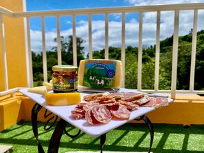 Ett bord med ost, en burk och skivad chark på en balkong med ett räcke och grönska i bakgrunden.