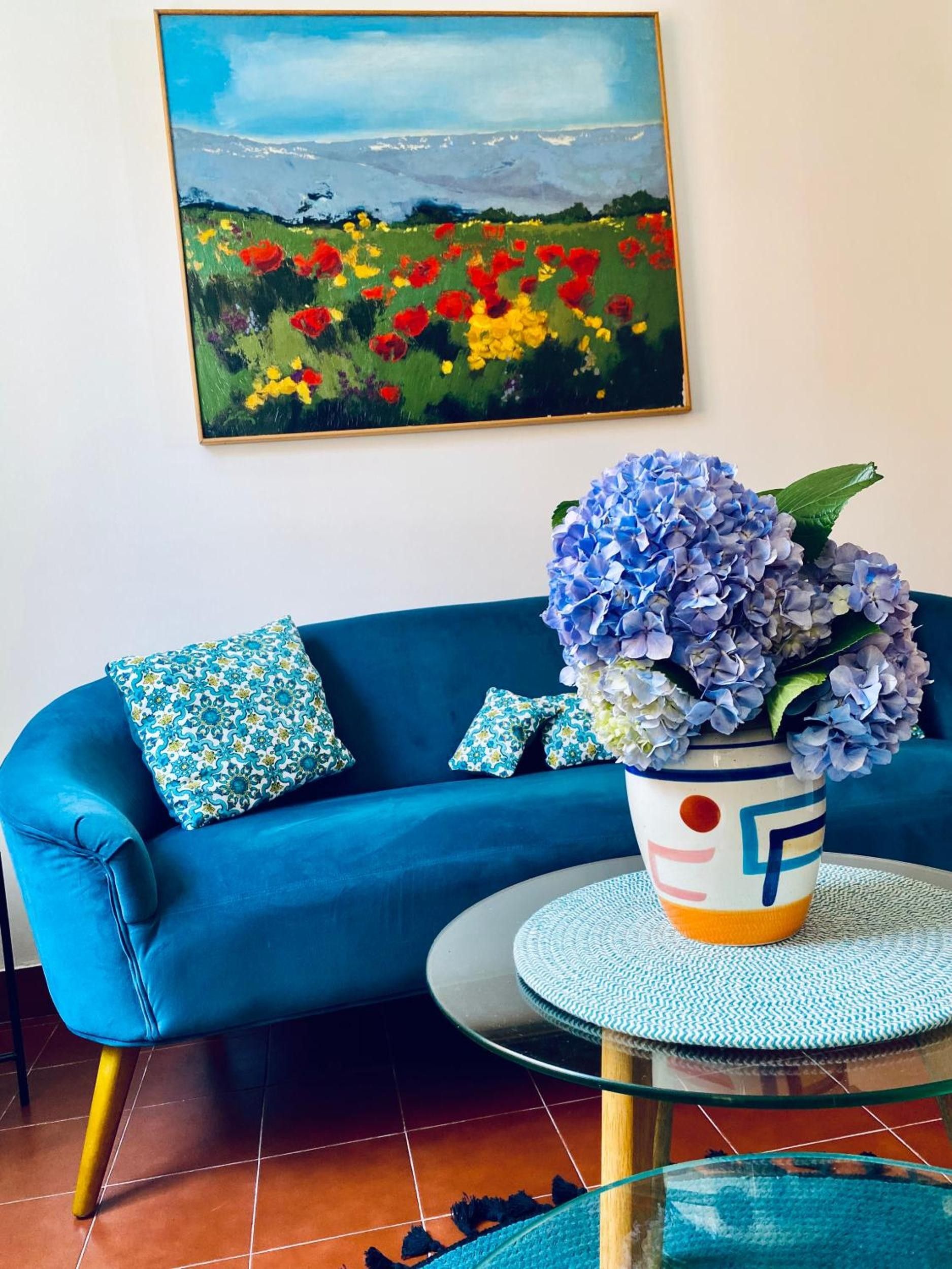 A blue sofa with patterned cushions, a glass coffee table, and a vase with blue flowers.