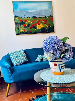Un divano blu con cuscini decorati, un tavolino in vetro e un vaso con fiori blu.