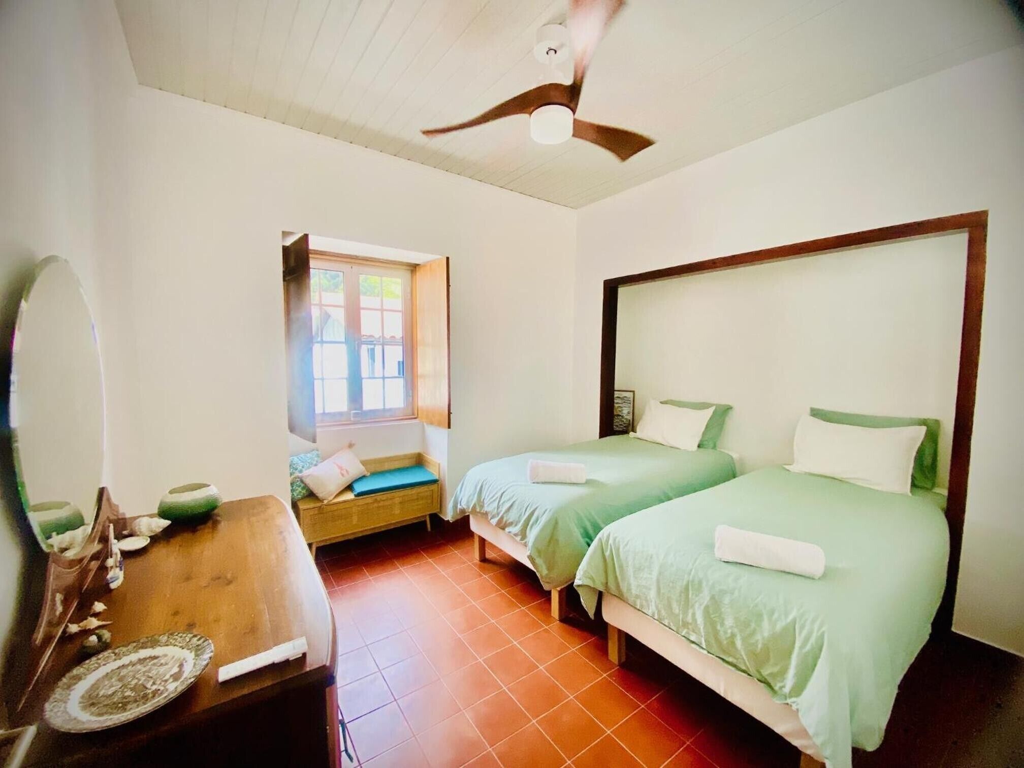 A bedroom with two beds, a wooden desk, a window, and a ceiling fan.