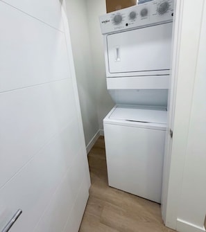 A compact laundry room with a stacked washer and dryer, a small shelf, and a door leading to another room. - New* Black Mountain Cozy Modern 2bedroom Private Suite (Kelowna)