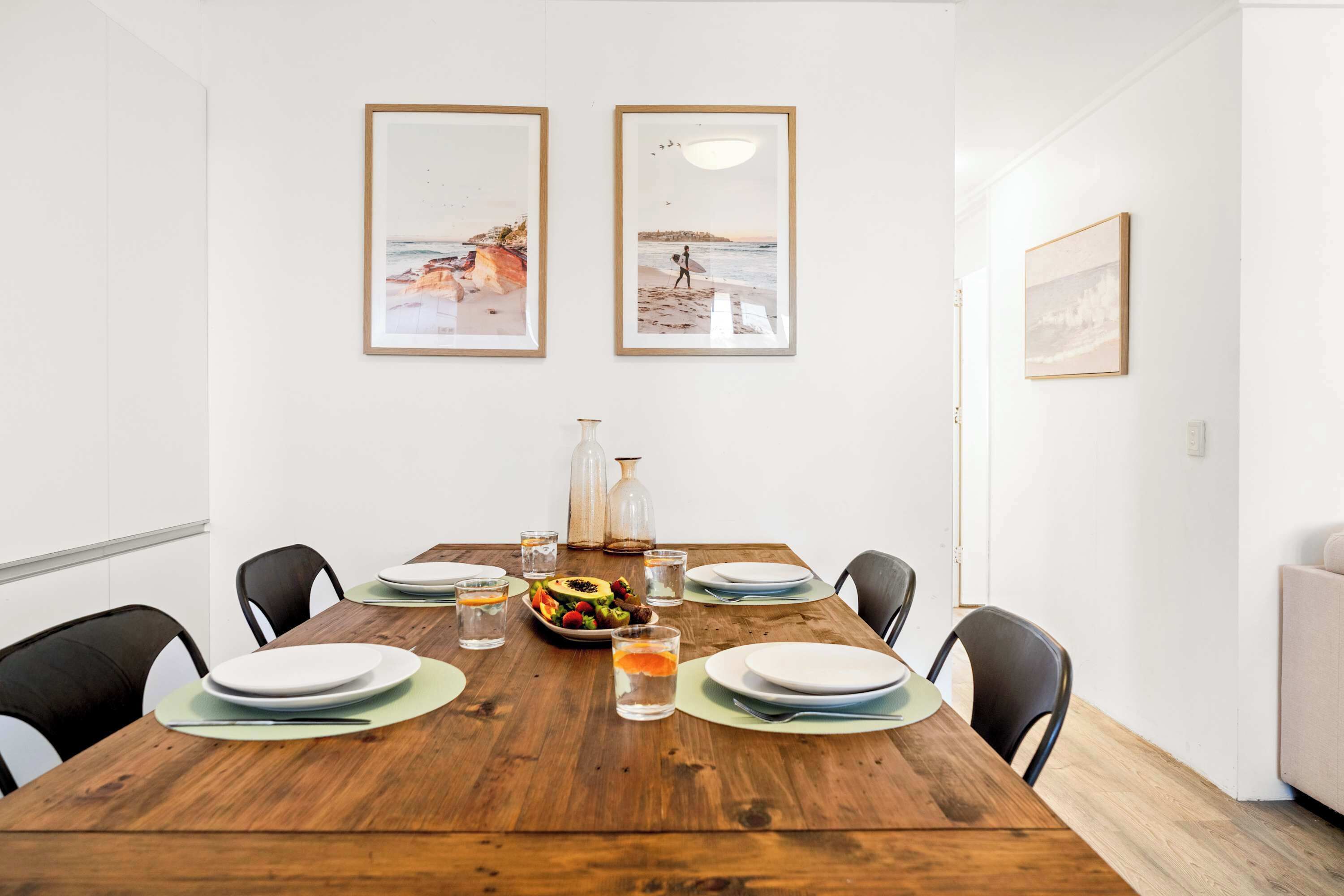 A dining area with a wooden table set for four, surrounded by chairs. There are framed pictures on the wall, a vase, and a bottle on the table.