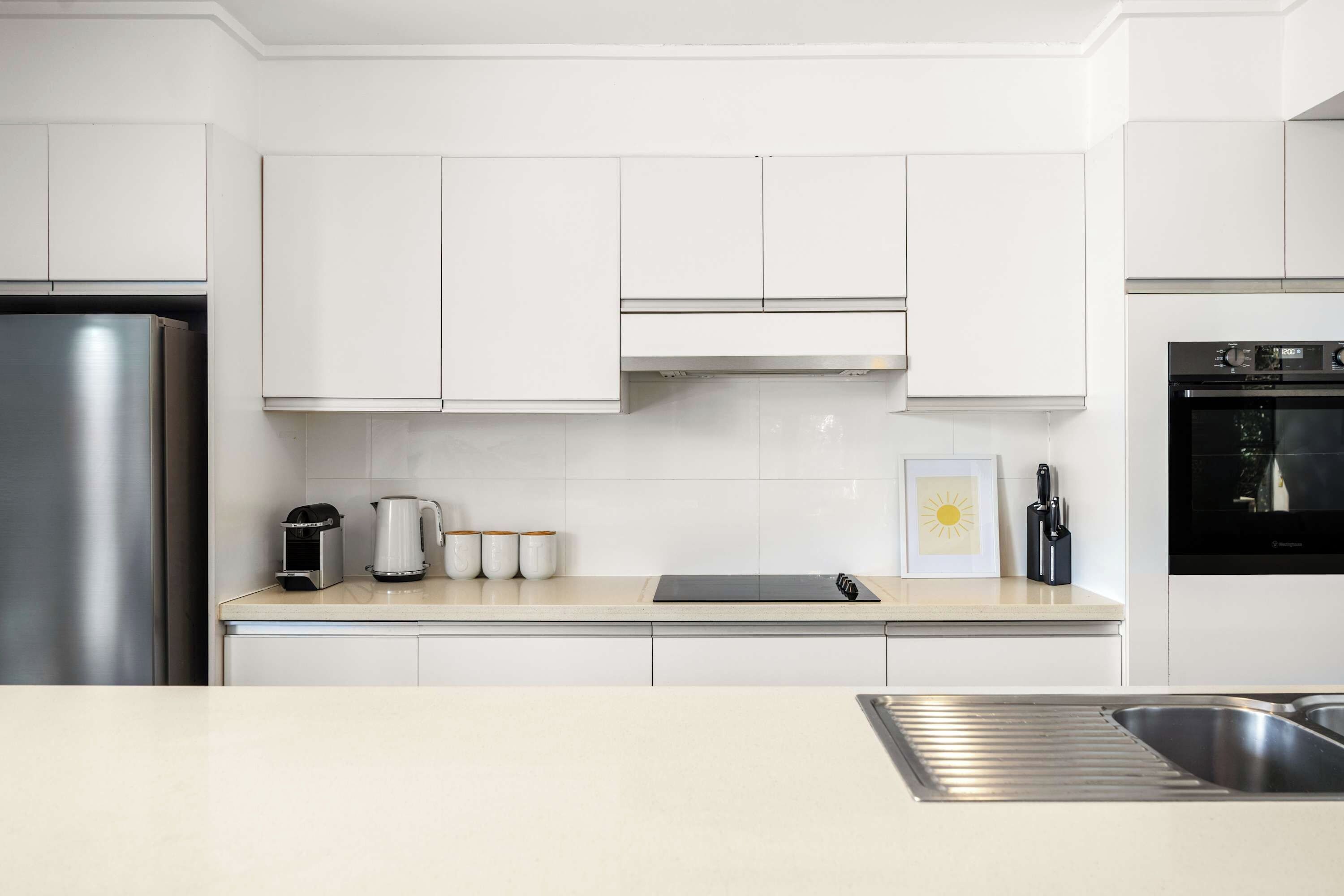 Eine moderne Küche mit weißen Schränken, einem Edelstahl-Kühlschrank, einer Kaffeemaschine und einem Wasserkocher auf der Arbeitsfläche.