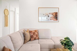 A living room with a beige sectional sofa, a framed landscape painting, a macrame wall hanging, and a potted plant. - Point Arkwright Escape - Rooftop, Pool & Views (Coolum Region)