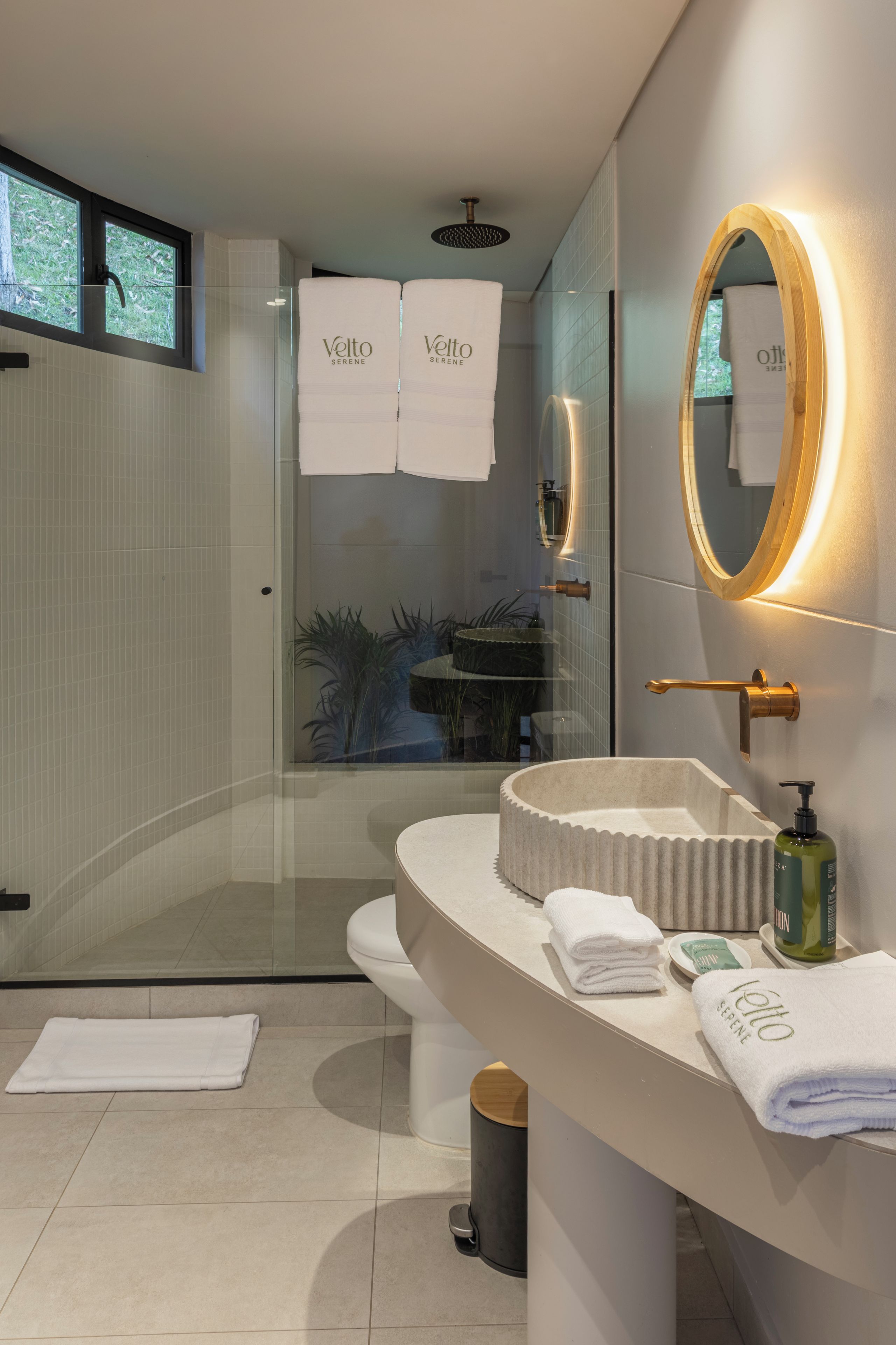 Un baño moderno con cabina de ducha de vidrio, un lavabo redondo con encimera color beige y un espejo grande con marco dorado.
