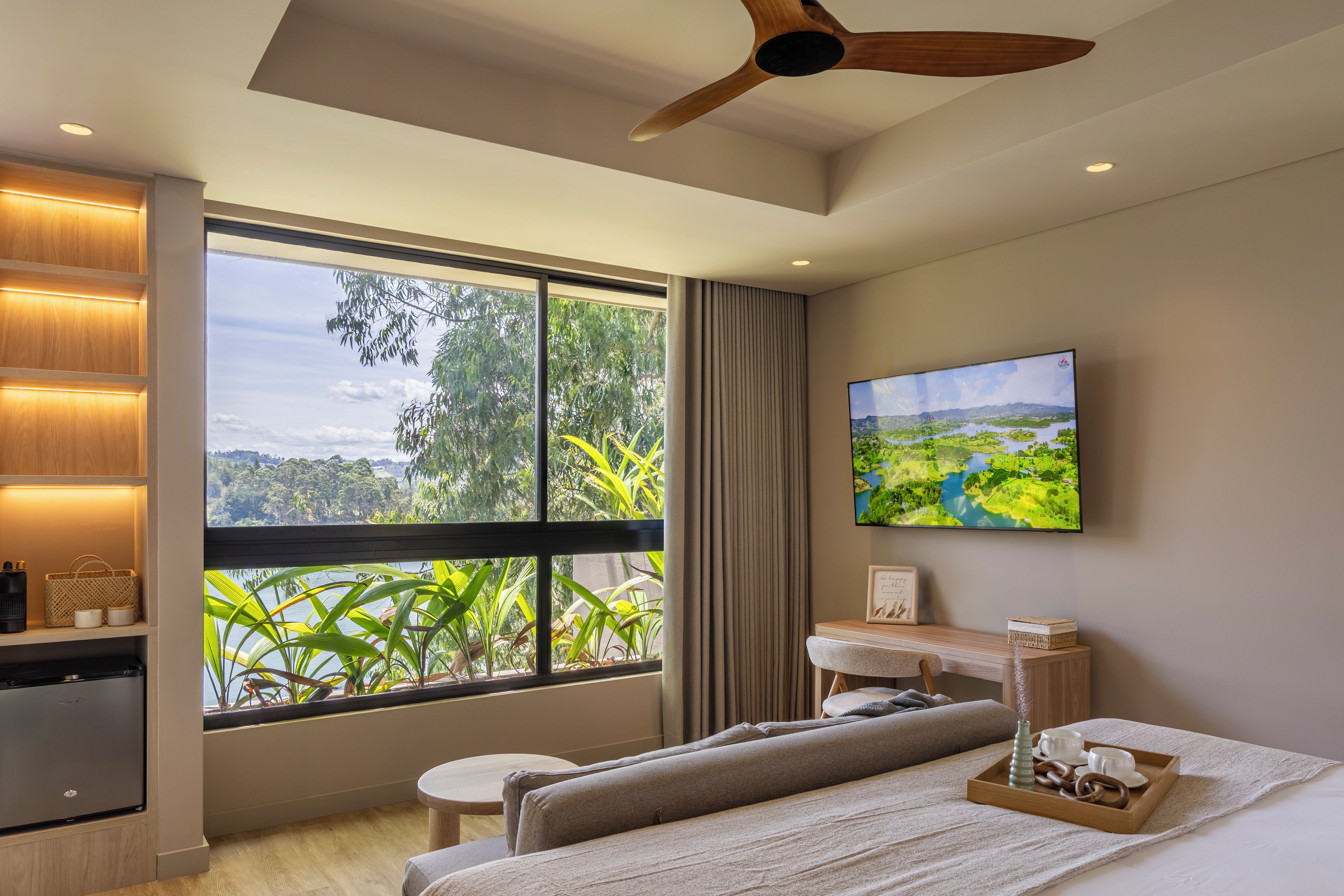Un dormitorio moderno con un ventanal grande, un ventilador de techo, un televisor de pantalla plana y una mesita de noche con una bandeja.