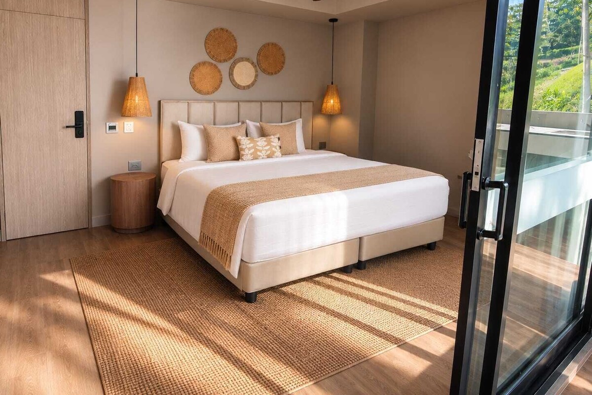 A modern bedroom with a large bed, a ceiling fan, and a view of greenery through a sliding door.