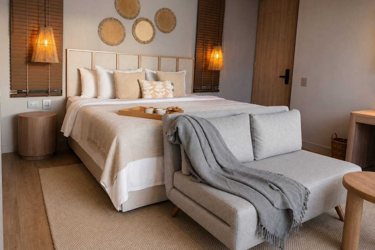 A bedroom with a bed, bedside lamps, a bedside table, a sofa, and a wall-mounted artwork.