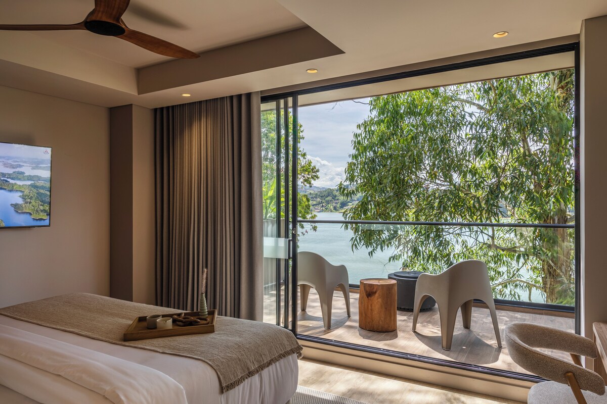 Habitación de hotel moderna con un ventanal grande, una cama, ventilador de techo y vistas a un lago.