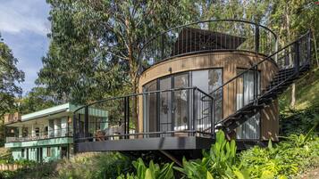 Una estructura moderna y circular de madera con cerramiento de vidrio, conectada a un edificio por una escalera de metal.