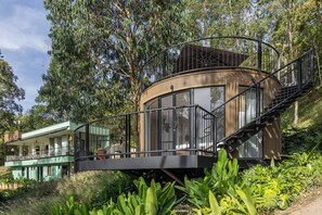 Una estructura moderna y circular de madera con cerramiento de vidrio, conectada a un edificio por una escalera de metal.