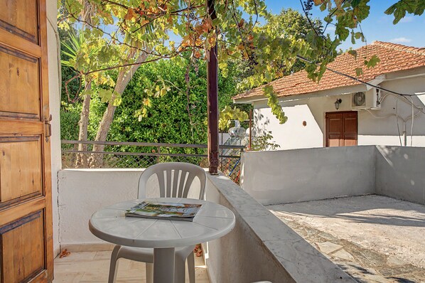 A balcony with a table and chairs, a wooden door, and a building with a tiled roof in the background. - Room 'Orama 25' with Sea View, Wi-Fi and Air Conditioning (Εύβοια)