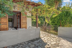 A stone-paved patio with a wooden door and shutters, surrounded by greenery and a metal fence. - Room 'Orama 25' with Sea View, Wi-Fi and Air Conditioning (Εύβοια)