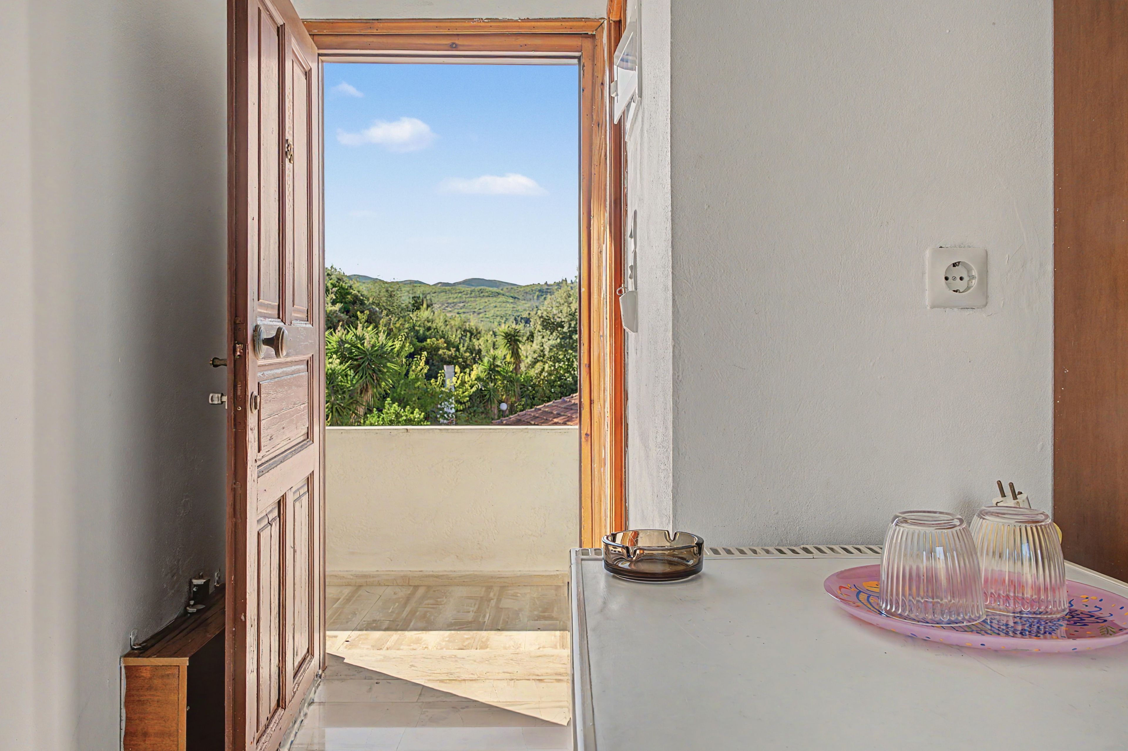 Ein Zimmer mit einer Holztür, die auf einen Balkon mit Blick auf Grünflächen und Hügel führt.