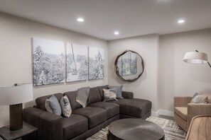 Een moderne woonkamer met een hoekbank, een rond bijzettafeltje en een salontafel. Drie aan de muur bevestigde kunstwerken tonen besneeuwde landschappen.