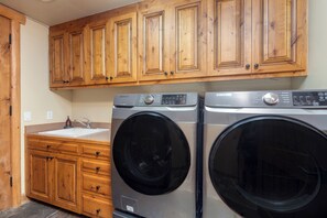 A laundry room with two front-loading washing machines, wooden cabinets above, and a sink with a faucet. - Luxury Mountain Village Retreat | Panoramic Mountain Views | Private Hot Tub | Sleeps 9 (Telluride)