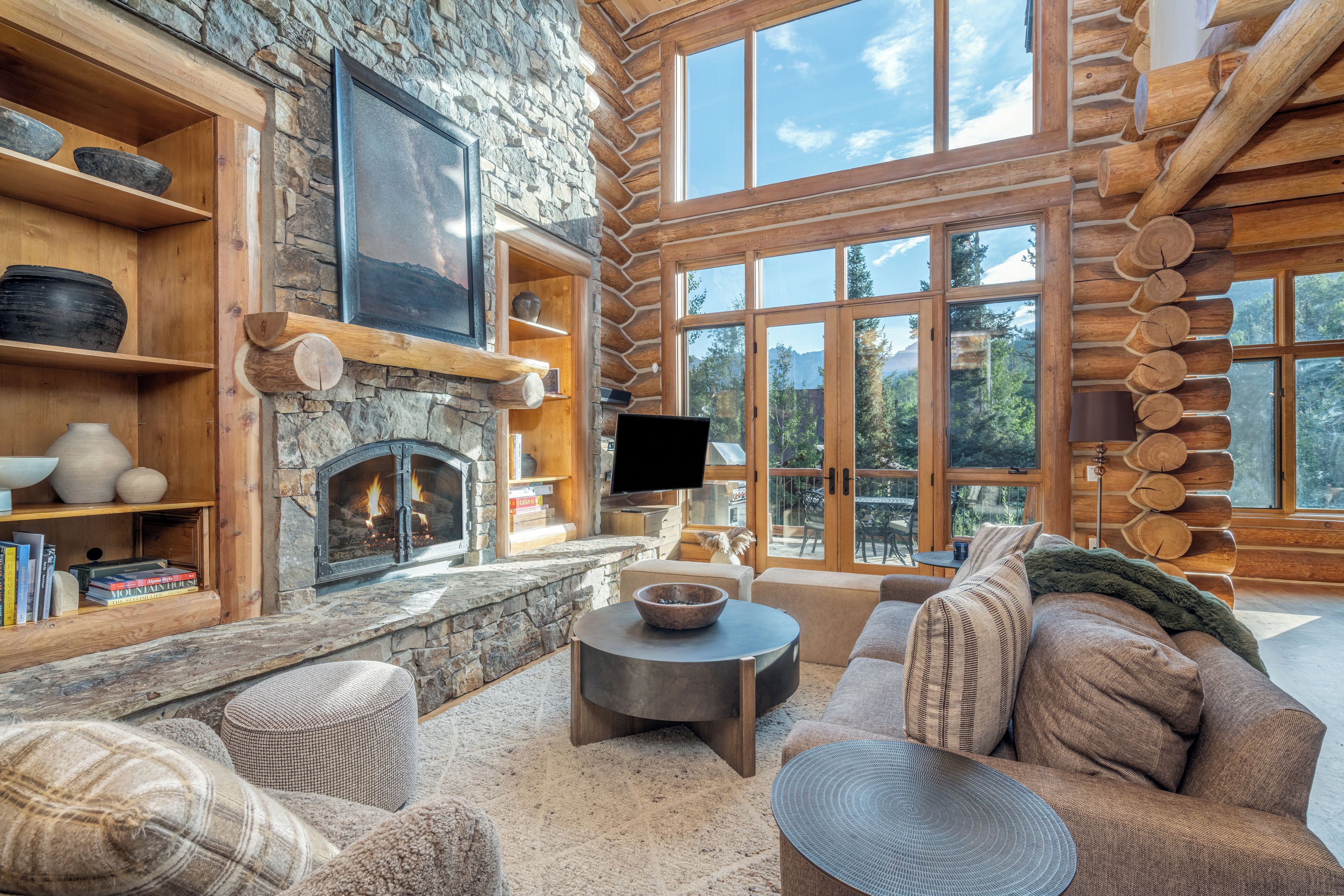 A log cabin interior with a stone fireplace, wooden shelves, a television, and a cozy seating area.