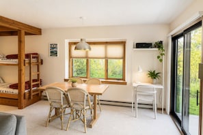 A room with a wooden bunk bed, a dining table with chairs, a hanging light fixture, a window with blinds, and a white desk with a lamp and plant. - JHRL - Elderberry 4011 (Wilson)