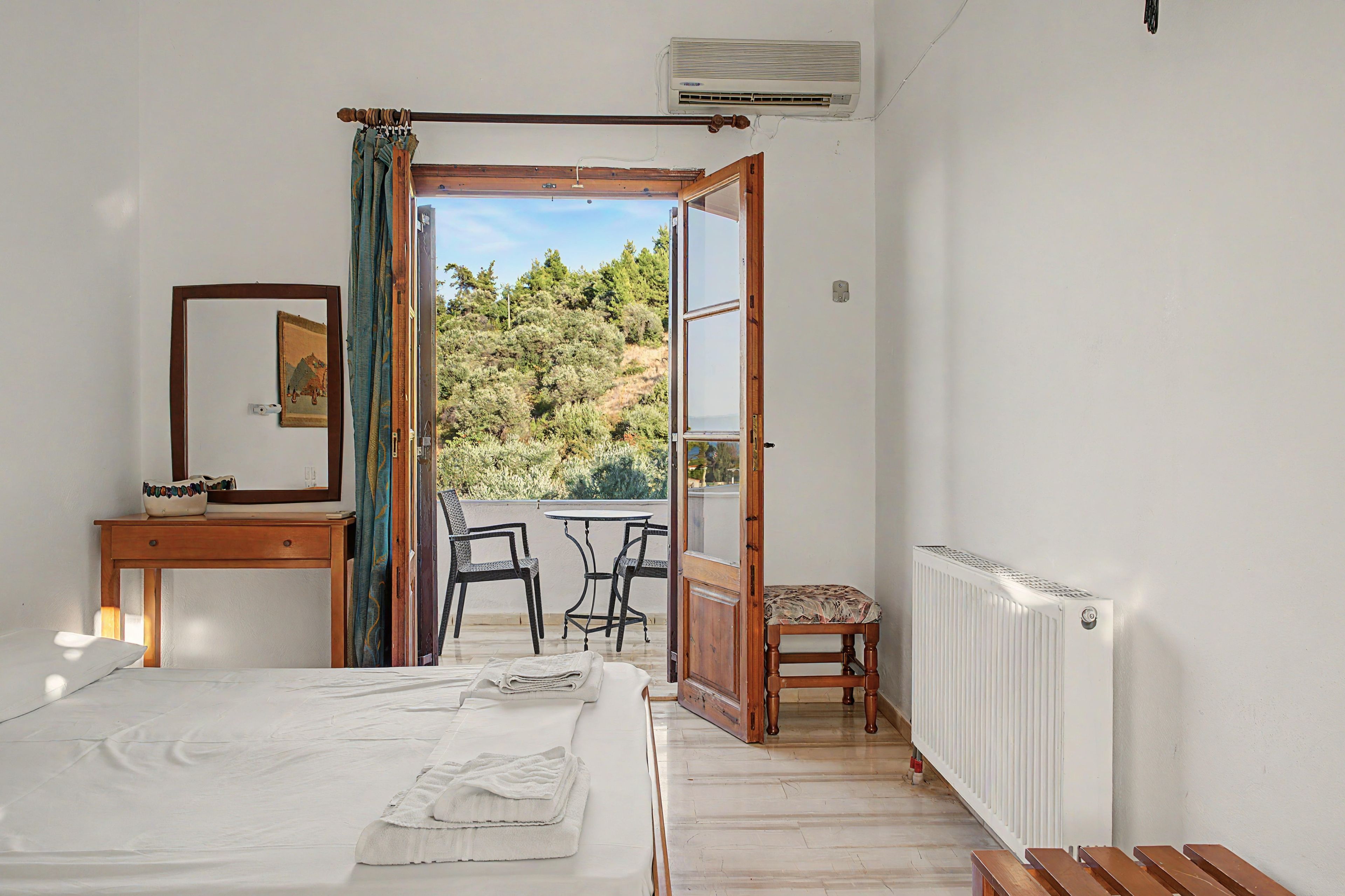 Ein Zimmer mit einem Bett, einem kleinen Tisch, einem Sessel, einer Kommode und einer offenen Tür, die auf einen Balkon mit Blick auf Bäume führt.