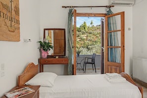 Ein Schlafzimmer mit einem Bett, einer Kommode, einem Spiegel, einer Zimmerpflanze und Blick ins Freie durch eine offene Tür.